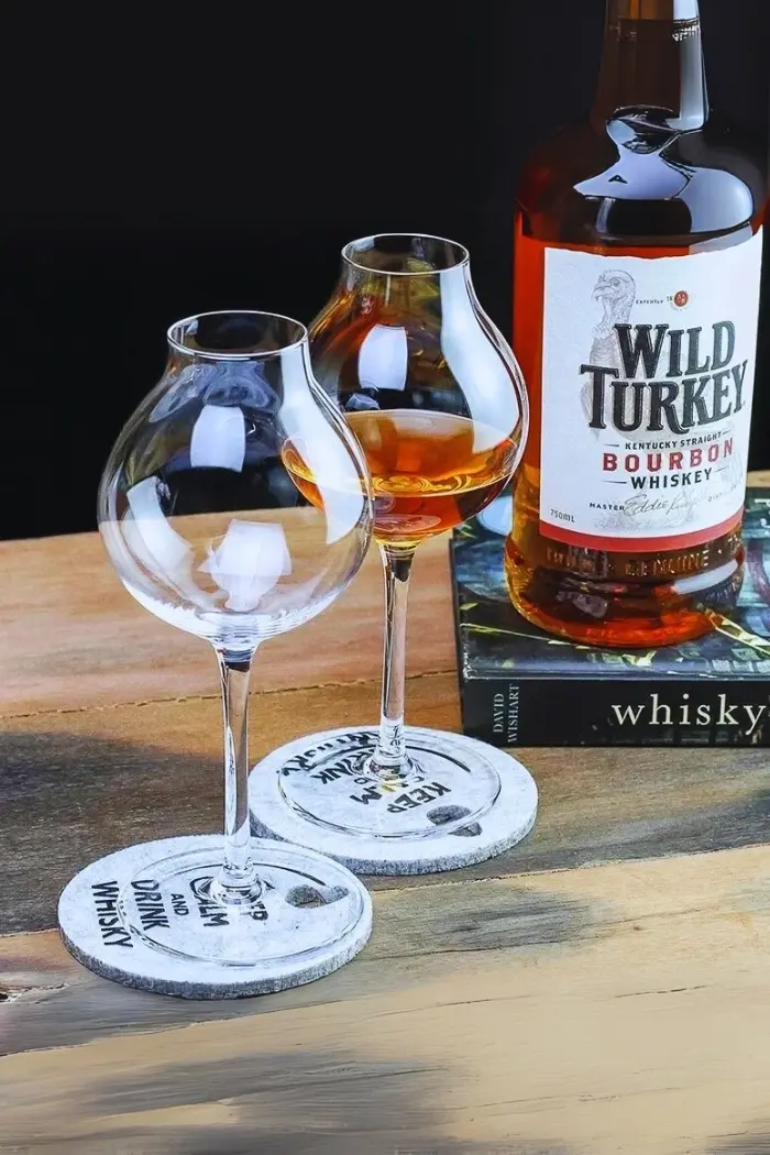 Verre à whisky tulipe posé sur sous-verre en pierre, avec bouteille de bourbon Wild Turkey en arrière-plan.