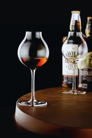 Verre à whisky tulipe rempli, posé sur table ronde avec bouteilles en arrière-plan sur fond noir.