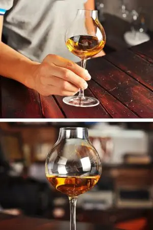 Verre à whisky tulipe tenu par la base, rempli, posé sur table foncée.