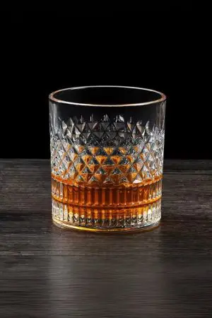 Verre à whisky cristal tumbler posé sur une surface en bois foncé, rempli partiellement de whisky ambré.