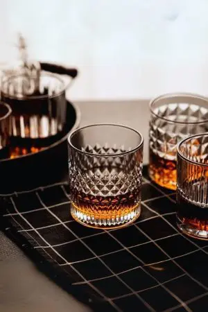 Verre à whisky cristal tumbler posé sur une nappe noire à motifs quadrillés, entouré d’autres verres partiellement remplis.