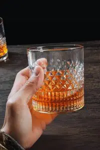 Verre à whisky cristal tumbler tenu en main au-dessus d'une table en bois foncé, rempli de whisky.