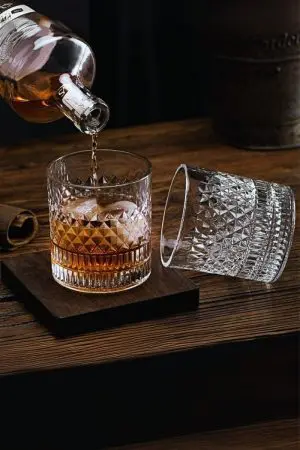 Verre à whisky cristal tumbler en cours de remplissage sur une planche en bois, avec un second verre vide couché à côté.