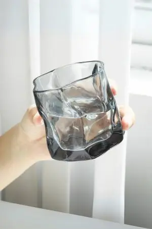 verre à whisky gris fumé incliné dans une main devant rideau blanc