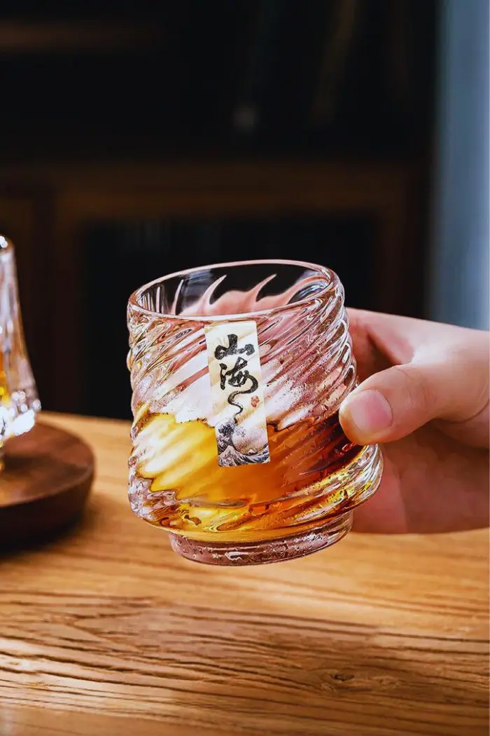 Verre à whisky rempli, dans la main, glaçons visibles et motif marin à la base