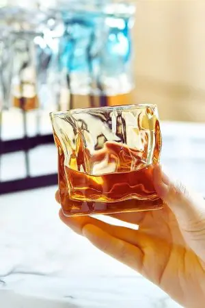 Verre à whisky tumbler ambre tenu en main devant support à verres.