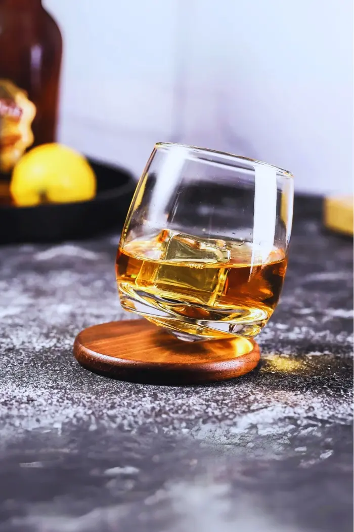 Verre à whisky tumbler incliné sur socle en bois rond, avec glaçon cubique.
