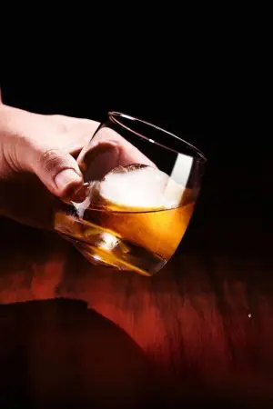 Verre à whisky tumbler tenu en main, avec glaçon sphérique et fond noir.