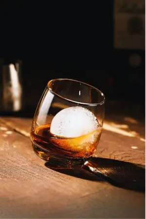Verre à whisky tumbler incliné avec glace sphérique sur table en bois sombre.