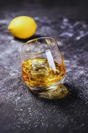 Verre à whisky tumbler incliné avec glaçon cubique sur surface grise texturée.