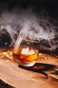 Verre à whisky tumbler incliné avec glaçon sur table en bois, dans une ambiance enfumée.