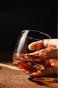 Verre à whisky tumbler tenu en main au-dessus d’une table en bois.