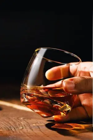 Verre à whisky tumbler tenu en main au-dessus d’une table en bois.