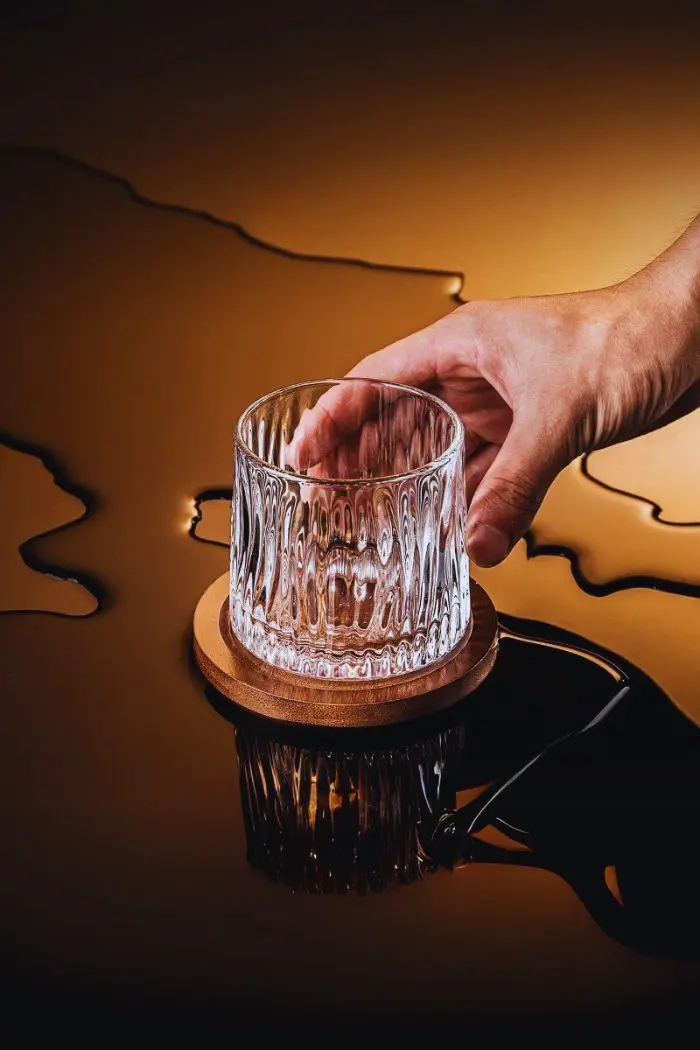 Verre à whisky tumbler vide, tenu par une main et posé sur un socle en bois foncé