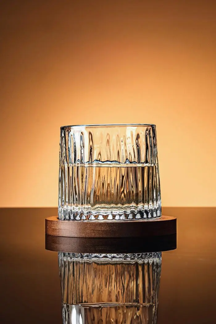 Verre à whisky tumbler rempli posé sur socle en bois avec fond orange.