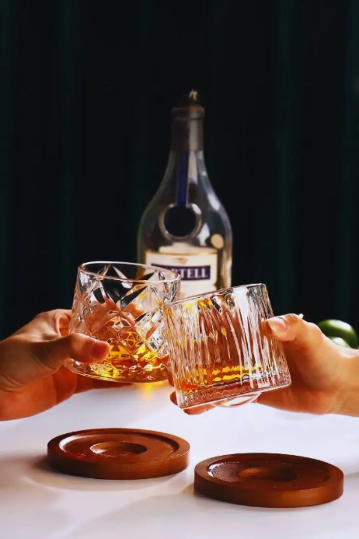 Verre à whisky tumbler texturé rempli, tenu à la main pour trinquer avec une bouteille en arrière-plan.