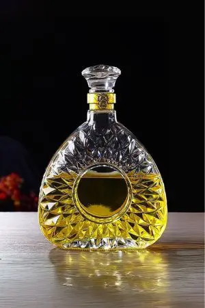 carafe à whisky ronde en verre taillé, bouchon avec bague dorée, remplie de whisky ambré, posée devant une fenêtre