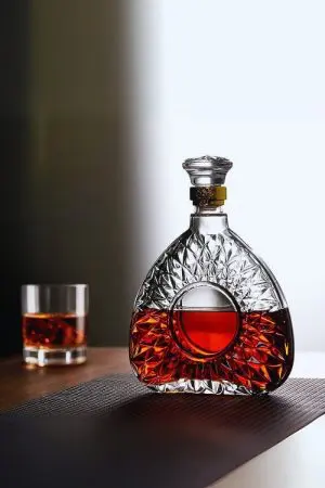 carafe à whisky ronde en verre taillé avec bague dorée, remplie de whisky foncé, posée sur une table en bois