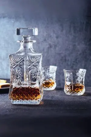carafe à whisky en verre taillé, partiellement remplie, accompagnée de deux verres assortis sur fond gris