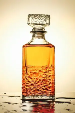 carafe à whisky carrée en verre transparent avec bouchon sculpté, posée sur une surface mouillée, remplie de whisky ambré