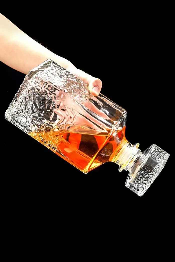 carafe à whisky carrée en verre tenue inclinée à la main, fond noir, remplie de whisky ambré