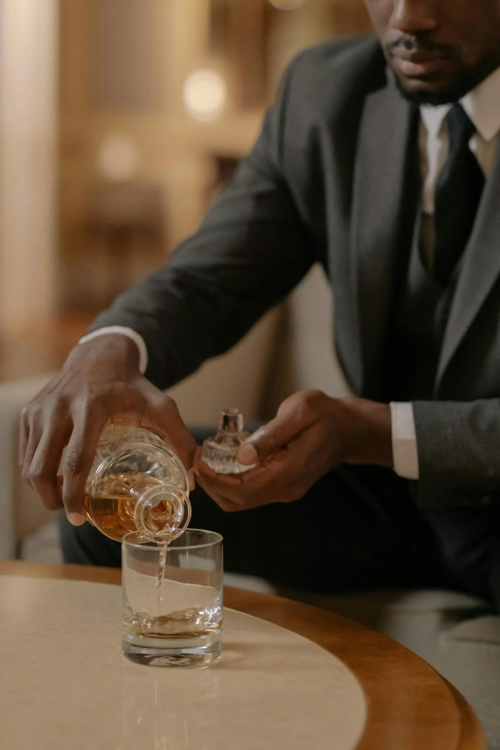 Un homme élégant sert du whisky à la main depuis une carafe en verre dans un verre tumbler, sur une table basse.