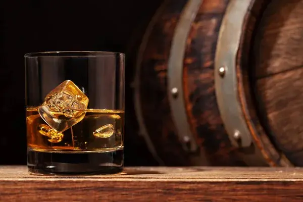 verre de whisky ambré avec glaçons posé devant des fûts en chêne