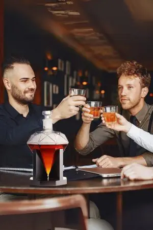 carafe à whisky diamant sur socle noir au centre d’une table, personnes trinquant