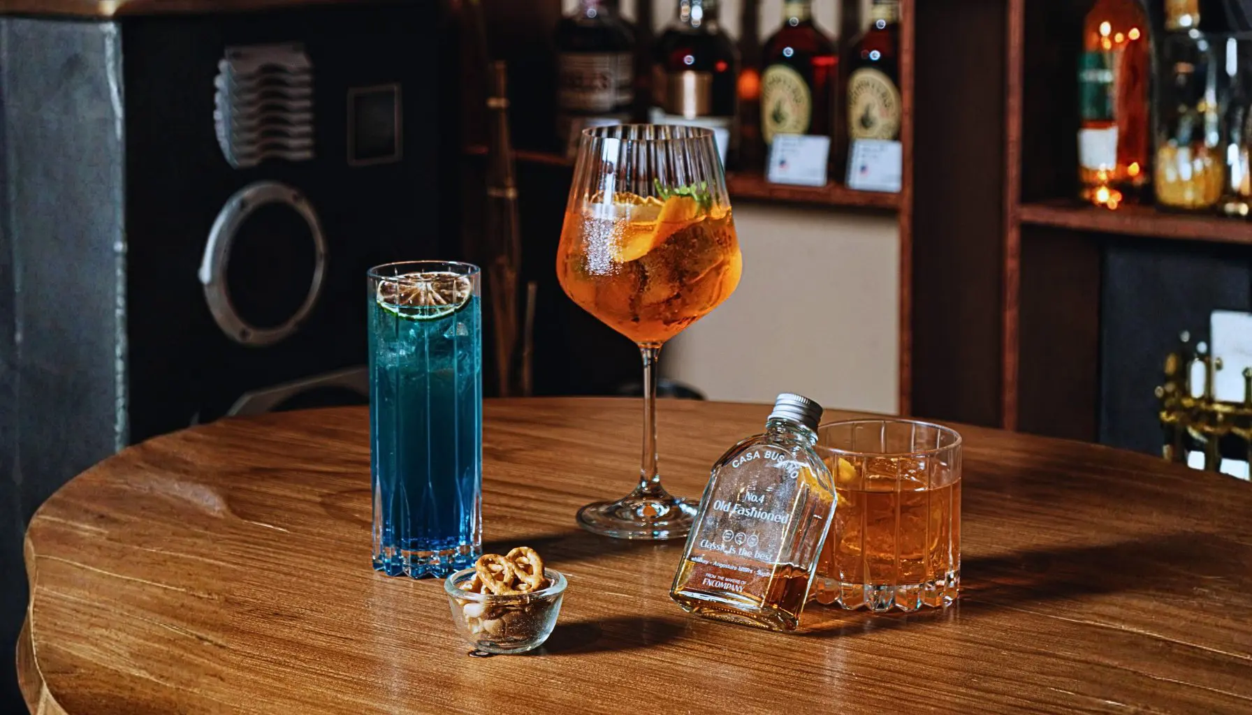 Différents verres et cocktails posés sur une table en bois dans un bar, montrant l’impact du choix du verre sur l’expérience de dégustation du whisky.