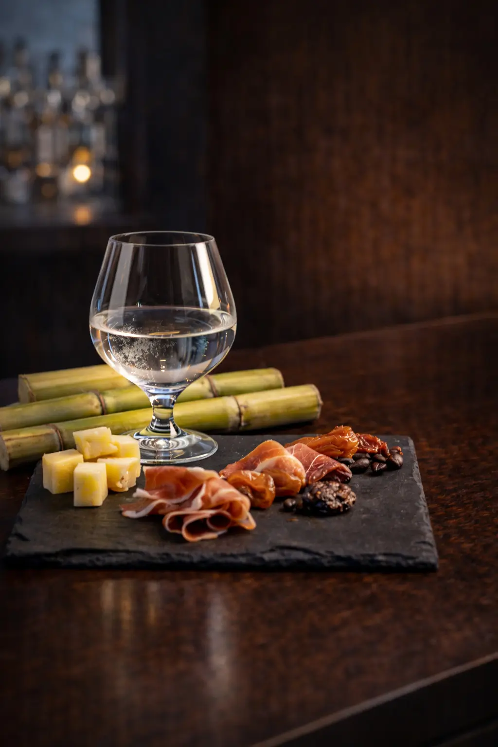 Un verre snifter contenant un rhum blanc clair posé sur un comptoir de bar en bois sombre, derrière une ardoise de dégustation avec des morceaux de fromage, du jambon cru, des dattes et des grains de café, le tout reposant sur des cannes à sucre.