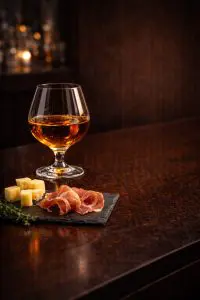 verre de rhum ambré accompagné de cubes de fromage affiné et de jambon cru sur ardoise