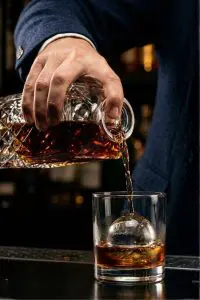 Gros plan d'une main versant du rhum depuis une carafe en cristal dans un verre old fashioned avec une sphère de glace.